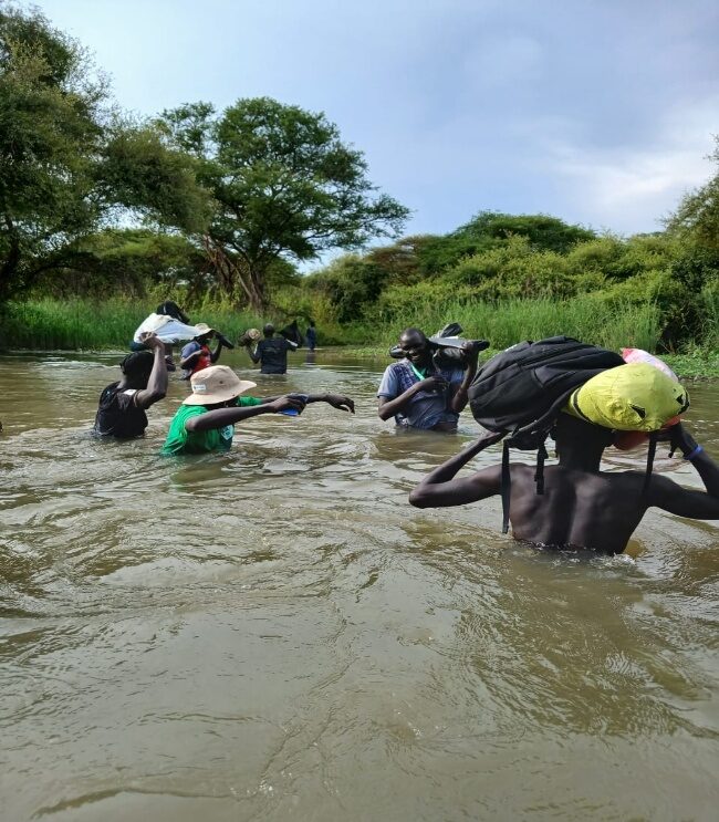 ARDF team members cross a deep stream on the way to communities in Uddier Payam, carrying supplies and equipment above the water.